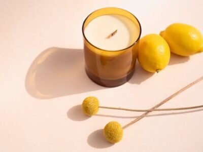 Scented candle in a brown glass holder beside lemons and yellow flowers on a white surface.